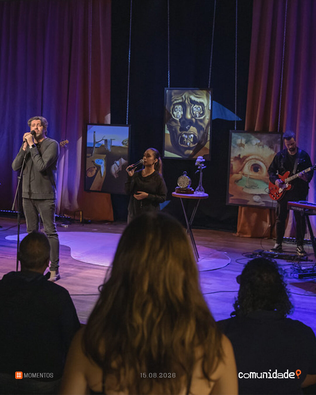 Palco do encontro com dois cantores ao microfone, o guitarrista e os quadros da série ao fundo, vistos por cima do público