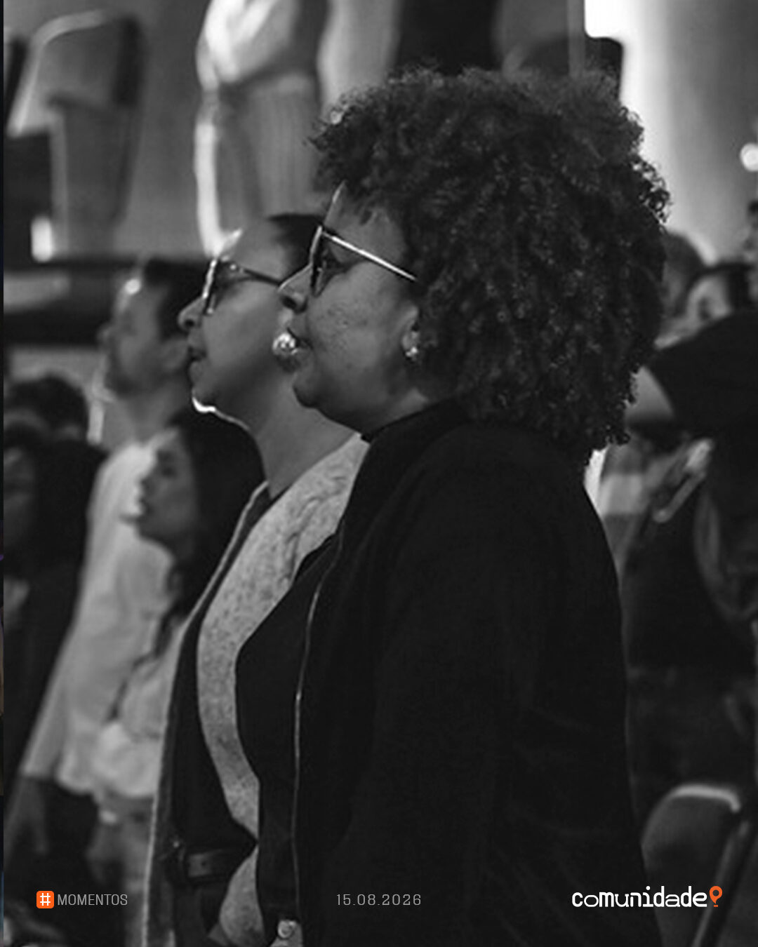 Em preto e branco, mulher de óculos cantando no meio da plateia em pé