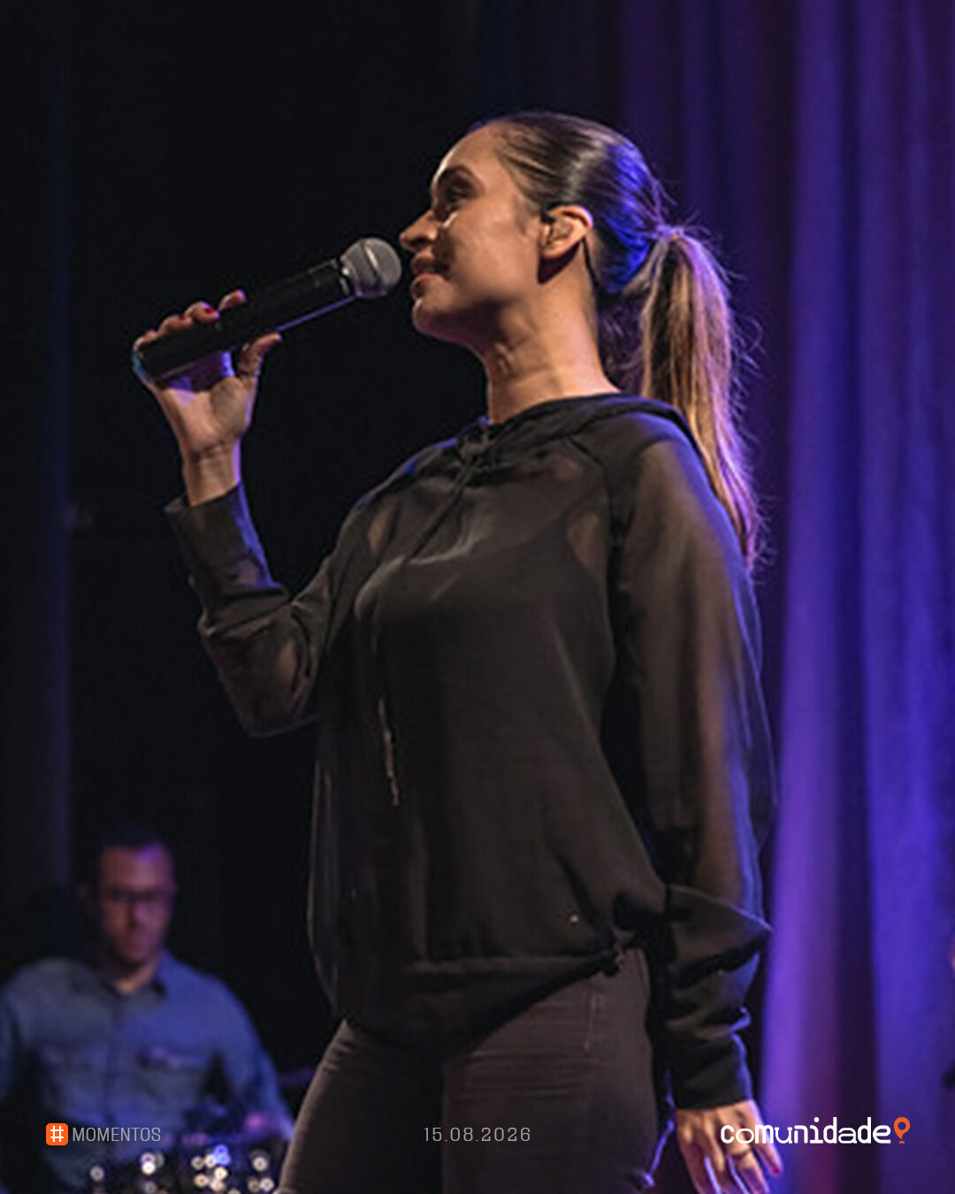 Cantora ao microfone no palco, de perfil, com a banda ao fundo